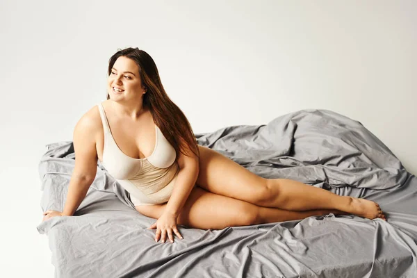 Charming woman with natural makeup and plus size body wearing beige bodysuit and posing on bed with grey bedding, body positive, figure type, smiling, looking away, diversity of body — Stock Photo