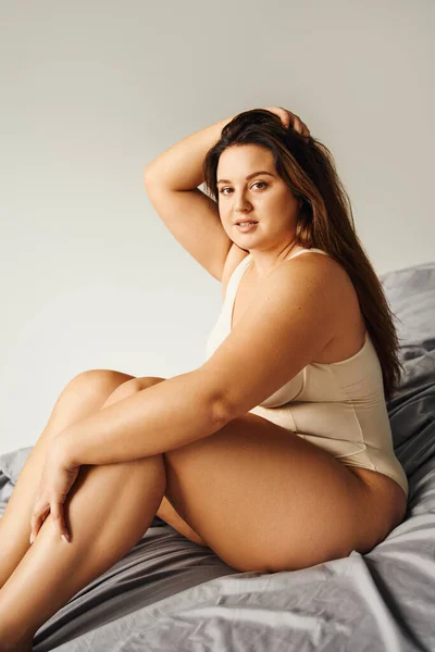 Brunette alluring woman with long hair and plus size body and bare feet wearing beige bodysuit and looking at camera while touching hair and sitting on bed with grey bedding, body positive — Stock Photo