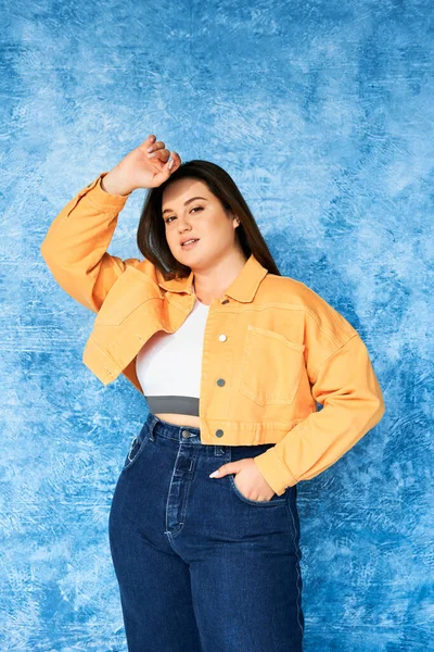 Plus size woman with long hair and natural makeup wearing crop top, orange jacket and denim jeans while posing with hand in pocket and looking at camera on mottled blue background, body positive — Stock Photo