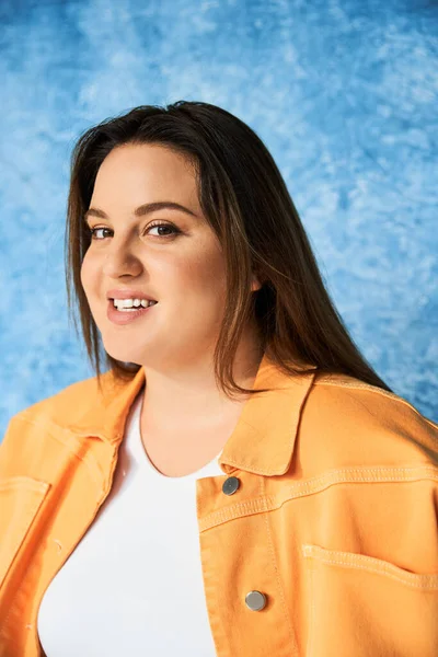 Portrait of positive plus size woman with long hair and natural makeup wearing crop top and orange jacket while posing and looking at camera on mottled blue background, body positive — Stock Photo