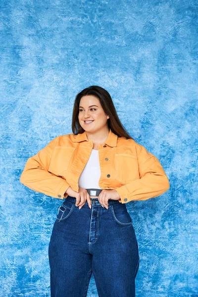 Positive plus size woman with long hair and natural makeup wearing crop top, orange jacket and adjusting denim jeans while posing and looking away on mottled blue background, body positive — Stock Photo