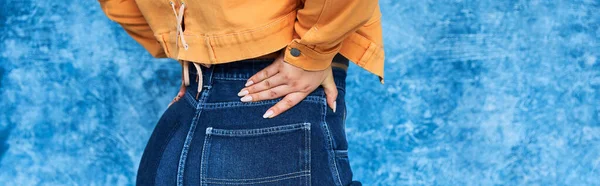 Vista recortada de mujer anónima de talla grande en chaqueta naranja y jeans de mezclilla posando con la mano en la cadera mientras está de pie con la mano en la cadera sobre fondo azul moteado, cuerpo positivo, pancarta - foto de stock