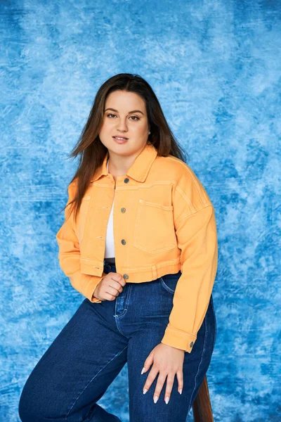 Brunette plus size woman with long hair and natural makeup wearing orange jacket and denim jeans while sitting on stool and looking at camera on mottled blue background, body positive — Stock Photo