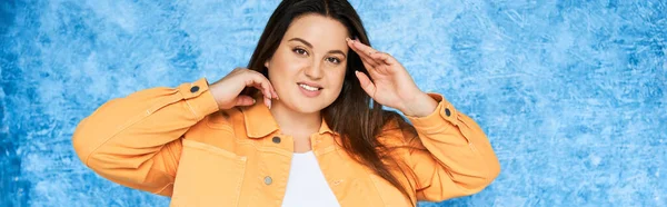 Self Acceptance, body positive, happy plus size woman with long hair and natural makeup wearing orange jacket while posing and looking at camera on mottled blue background, banner — Stock Photo