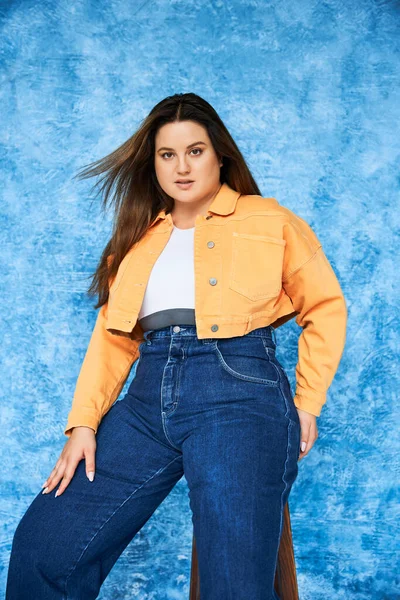 Позитивная по телу и брюнетка plus-size женщина с длинными волосами и естественным макияжем в топе, оранжевой куртке и джинсах с джинсами, позируя и глядя в камеру на синем фоне — стоковое фото