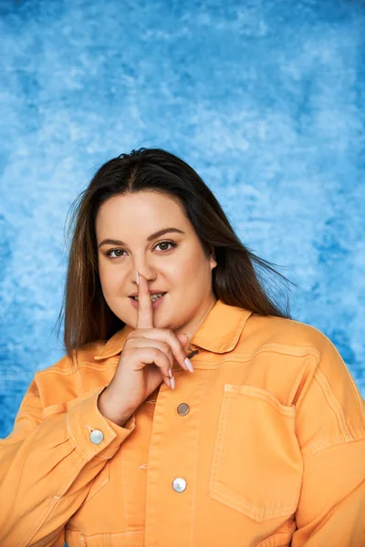 Body positive and brunette plus size woman with brunette hair and natural makeup wearing orange jacket and showing hush sign and looking at camera on mottled blue background — Stock Photo