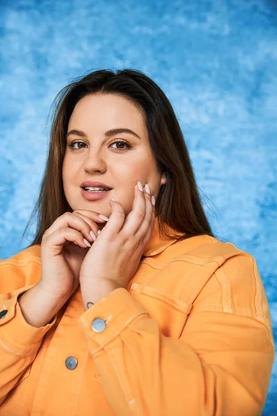 Portrait of body positive and brunette plus size woman with long hair and natural makeup wearing orange jacket, touching face and looking at camera on mottled blue background — Stock Photo