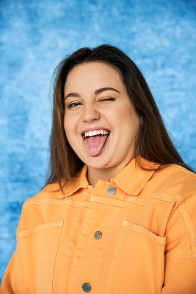 Portrait of funny plus size woman with brunette hair and natural makeup wearing orange jacket and sticking out tongue while looking at camera on mottled blue background, body positive — Stock Photo