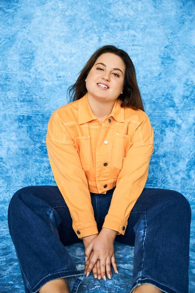 Позитивная по телу и брюнетка plus size женщина с естественным макияжем в оранжевой куртке и джинсах, позируя и глядя в камеру на синем фоне, счастливое лицо — стоковое фото