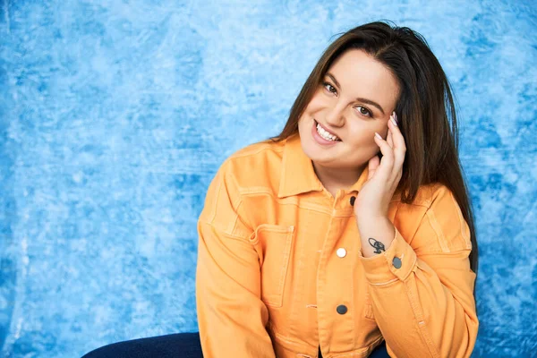 Tattooed and smiling plus size woman with brunette hair and natural makeup wearing orange jacket and touching cheek while posing and looking at camera on mottled blue background, body positive — Stock Photo