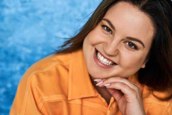 Portrait of joyous plus size woman with brunette hair and natural makeup wearing orange jacket and touching face while posing and looking at camera on mottled blue background, body positive — Stock Photo
