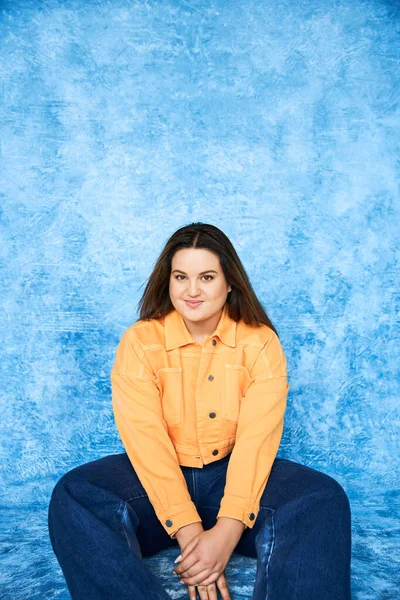 Body positive, plus size woman with brunette hair and natural makeup sitting in orange jacket and denim jeans while smiling and looking at camera on mottled blue background — Stock Photo