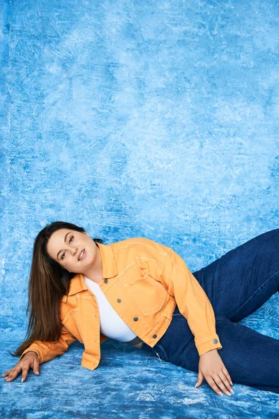 Mujer de tamaño grande con cabello moreno y maquillaje natural usando top de la cosecha, chaqueta naranja y jeans de mezclilla mientras posando y mirando a la cámara sobre fondo azul moteado, cuerpo positivo - foto de stock