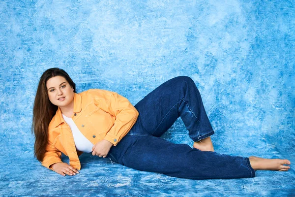 Повна довжина тіла позитивна і брюнетка плюс розмір жінки з довгим волоссям і натуральним макіяжем в помаранчевій куртці і джинсових джинсах, позуючи і дивлячись на камеру на строкатому синьому фоні — стокове фото