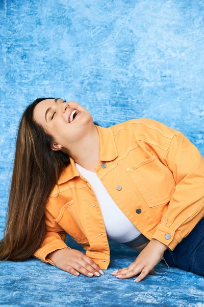 Body positive and happy plus size woman with long hair and natural makeup laughing with closed eyes while posing in orange jacket and denim jeans on mottled blue background — Stock Photo