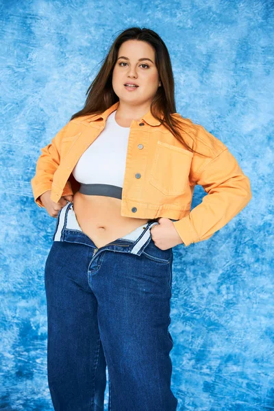 Позитивная по телу и брюнетка plus-size женщина с длинными волосами и естественным макияжем, в сером топе и оранжевой куртке в джинсах из джинсов, позирующая и смотрящая на камеру на темно-синем фоне — стоковое фото
