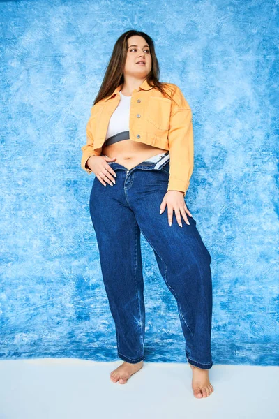Volle Länge der Barfuß-Körper positive Frau mit Plus-Size-Körper und brünetten Haaren posiert in orangefarbener Jacke, Crop Top und Jeans, während posiert und in die Kamera auf fleckigem blauem Hintergrund schaut — Stockfoto