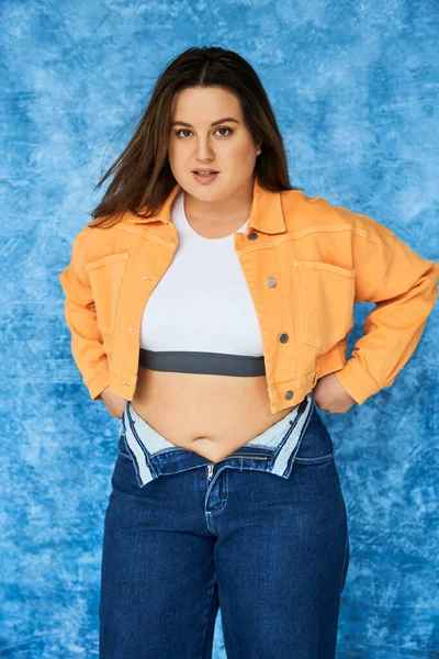 Body positive woman with long hair and natural makeup posing in crop top and orange jacket while wearing denim jeans while standing and looking at camera on mottled blue background — Stock Photo