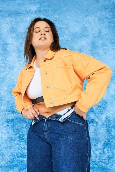Брюнетка тіло позитивна жінка з натуральним макіяжем і закритими очима позують у верхній і помаранчевій куртці, одягнені в джинсові джинси і стоять на строкатому синьому фоні, плюс розмір жінки — стокове фото
