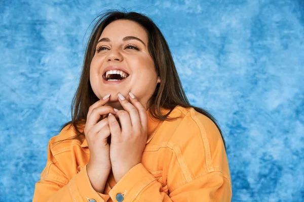 Portrait of body positive and happy plus size woman with brunette hair and natural makeup laughing while touching face and posing in orange jacket on mottled blue background — Stock Photo