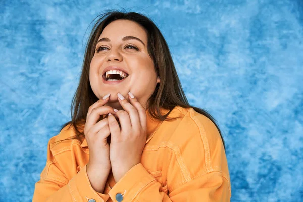 Retrato del cuerpo positivo y feliz mujer de talla grande con cabello moreno y maquillaje natural riendo mientras toca la cara y posando en chaqueta naranja sobre fondo azul moteado - foto de stock