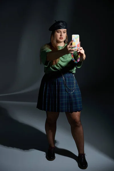 Plus size woman posing in leather beret, green t-shirt, plaid skirt with chains, fishnet tights and black shoes, taking selfie on smartphone on dark background, tattoo translation: harmony — Stock Photo