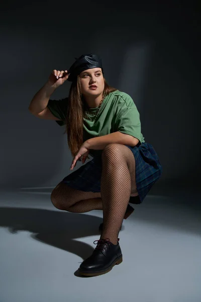Frau in voller Länge posiert in Lederbarett, grünem T-Shirt, kariertem Rock mit Ketten, Netzstrumpfhose und schwarzen Schuhen sitzend und wegschauend auf dunklem Hintergrund mit Studiobeleuchtung — Stockfoto