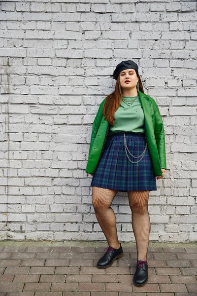 Full length of chic plus size woman posing in plaid skirt with chains, green leather jacket, beret, fishnet tights and black shoes while posing near brick wall on urban street, body positive — Stock Photo