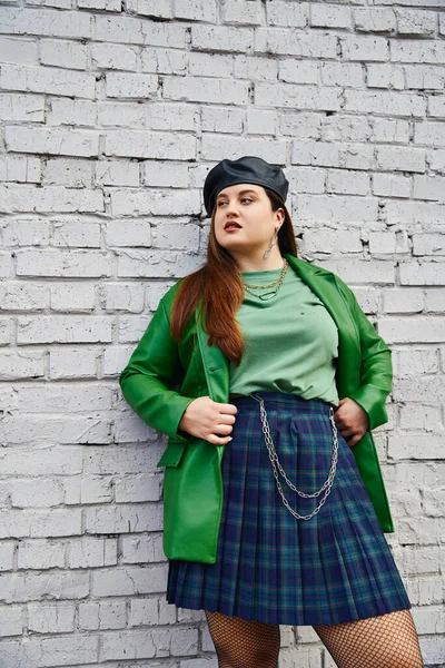 Chic plus taille femme posant en jupe à carreaux avec des chaînes, veste en cuir vert, béret noir et collants résille tout en regardant loin et debout près du mur de briques sur la rue urbaine, corps positif — Photo de stock