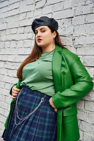 Stylish plus size woman posing in plaid skirt with chains, green leather jacket and black beret looking at camera and standing near brick wall on urban street, body positive — Stock Photo