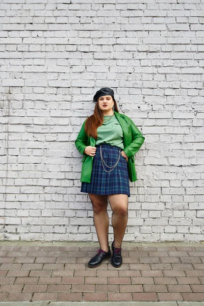 Full length of brunette plus size woman posing in leather jacket with beret, plaid skirt with chains, fishnet tights and black shoes while standing near brick wall on urban street — Stock Photo