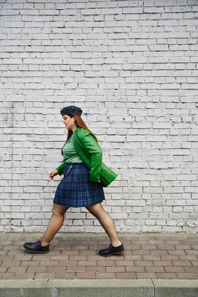 Full length of chic plus size woman posing in leather jacket, beret, plaid skirt with chains, fishnet tights and black shoes, walking near brick wall on urban street, body positive — Stock Photo