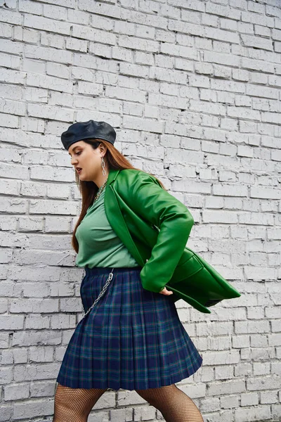 Seitenansicht der stylischen Plus-Size-Frau in grüner Lederjacke, schwarzer Baskenmütze, kariertem Rock mit Ketten und Netzstrumpfhose, die in der Nähe von Backsteinmauern auf urbaner Straße spaziert, körperbetont, selbstverliebt, urban chic — Stockfoto