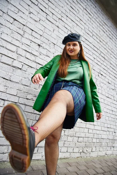 Low angle view of chic plus size woman posing in leather beret and jacket plaid skirt with chains, fishnet tights and black shoes while posing near brick wall on urban street, body positive — Stock Photo