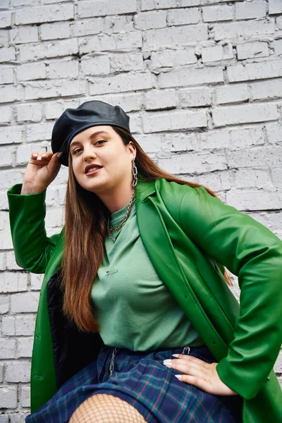 Stylish plus size woman in green leather jacket smiling while touching black beret and posing in plaid skirt and fishnet tights near brick wall on urban street, body positive, self-love, urban chic — Stock Photo