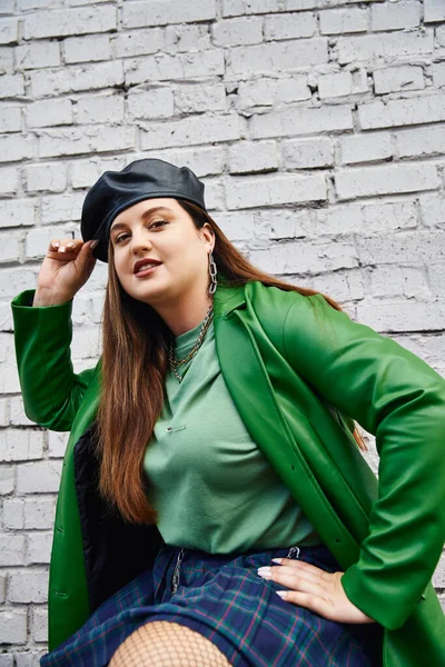 Elegante plus size donna in giacca di pelle verde sorridente mentre tocca berretto nero e posa in gonna a quadri e calze a rete vicino al muro di mattoni sulla strada urbana, corpo positivo, self-love, urban chic — Foto stock