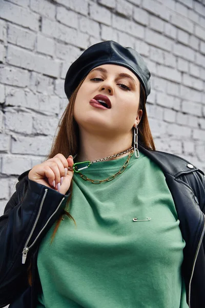 Stylish plus size woman in leather jacket and black beret pulling chain necklace while looking at camera and sticking tongue near brick wall on urban street, body positive, self-love, urban chic — Stock Photo