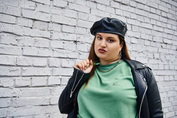 Stylish plus size woman in leather jacket and black beret pulling chain necklace while looking at camera near brick wall on urban street, body positive, self-love, unapologetic — Stock Photo