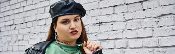 Stylish plus size woman in leather jacket and black beret pulling chain necklace while looking at camera near brick wall on urban street, body positive, self-love, unapologetic, banner — Stock Photo