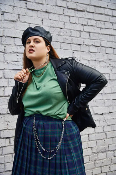 Stylish plus size woman in leather jacket and black beret pulling chain necklace while looking at camera and standing with hand on hip near brick wall on urban street, body positive, urban chic — Stock Photo