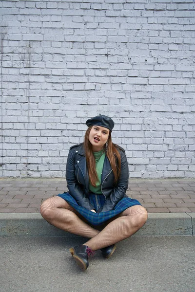 Full length of chic plus size woman posing in leather jacket, plaid skirt with chains, fishnet tights and black shoes while sitting near brick wall on urban street, body positive, full length — Stock Photo