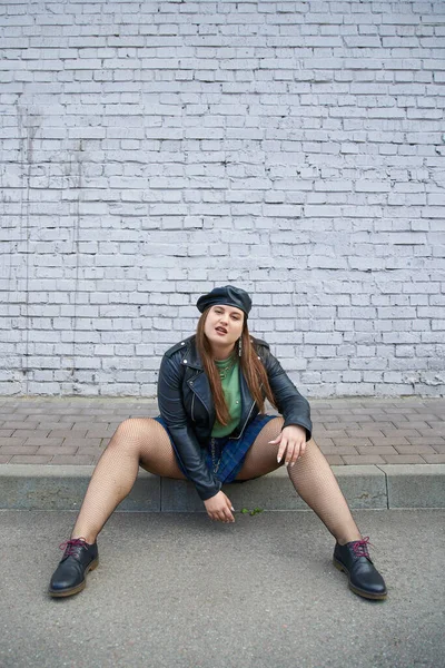 Fashionable plus size woman posing in leather jacket and beret, plaid skirt, fishnet tights and black shoes while posing near brick wall on urban street, body positive, full length — Stock Photo