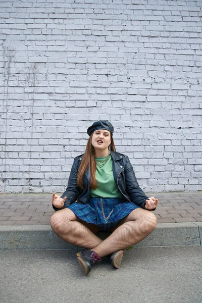 Chic plus size woman posing in leather beret and jacket, plaid skirt with chains, fishnet tights and black shoes while sitting with crossed legs near brick wall on urban street, rock sign — Stock Photo