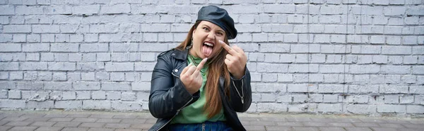 Sassy plus size woman sitting in leather jacket and beret while showing middle fingers and sticking tongue out near brick wall on urban street, body positive, bad behavior, banner — Stock Photo