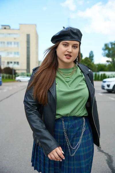 Brunette woman with plus size body walking in leather jacket with black beret, plaid skirt with chains and greet t-shirt near blurred building and parked car on urban street outdoors, body positive — Stock Photo
