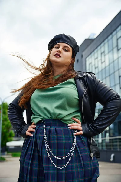 Confident woman with plus size body posing in leather jacket with black beret near blurred modern building on urban street outdoors, body positive, hands on hips, looking at camera — Stock Photo