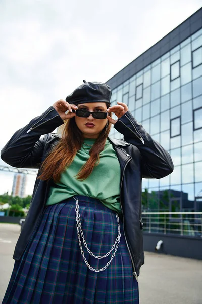 Low angle view of woman with plus size body standing in leather jacket with black beret, plaid skirt and greet t-shirt while wearing stylish sunglasses near blurred modern building on urban street — Stock Photo