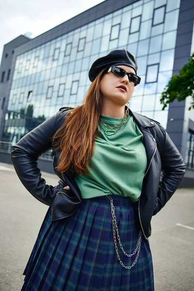 Brunette woman with plus size body posing with hands on hips while standing in stylish sunglasses, leather jacket with black beret, plaid skirt near blurred modern building on urban street — Stock Photo