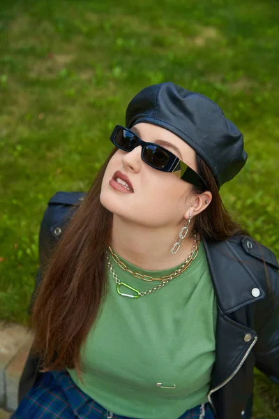 Vista de ángulo alto de mujer de talla grande en gafas de sol, chaqueta de cuero con boina negra y accesorios de cadena mirando hacia arriba en el parque verde con césped fresco, cuerpo positivo, al aire libre - foto de stock