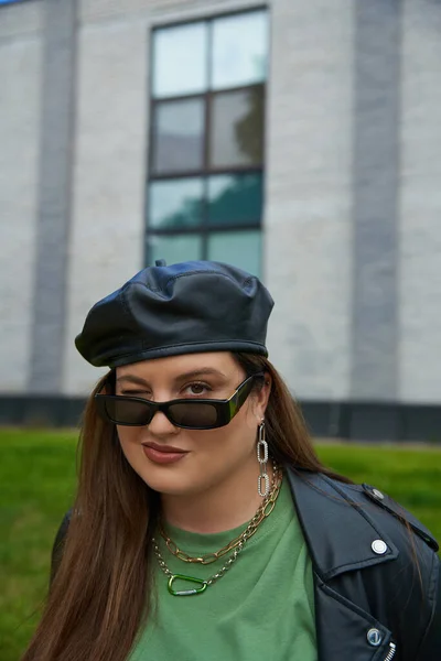 Porträt einer brünetten Frau in Plus-Size-Optik mit stylischer Sonnenbrille, Lederjacke mit schwarzem Baskenmütze und grüßendem T-Shirt, die in der Nähe eines verschwommenen modernen Gebäudes auf der städtischen Straße im Freien zwinkert, körperbetont — Stockfoto